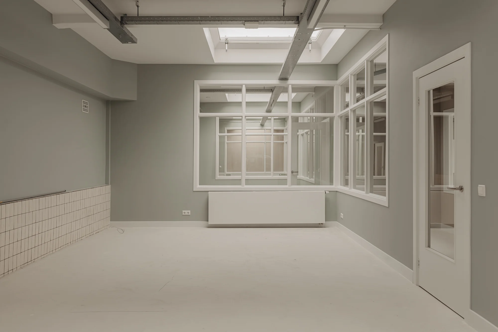 Lichtgrijze, lege kamer aan de Henri Polaklaan met grote ramen, plafondbalken en een glazen deur.